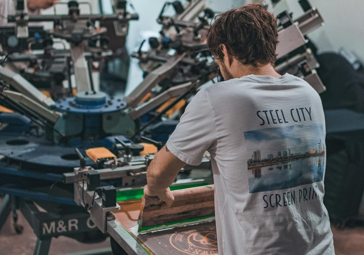 A man in a white t-shirt with a back print is working a screen printer.